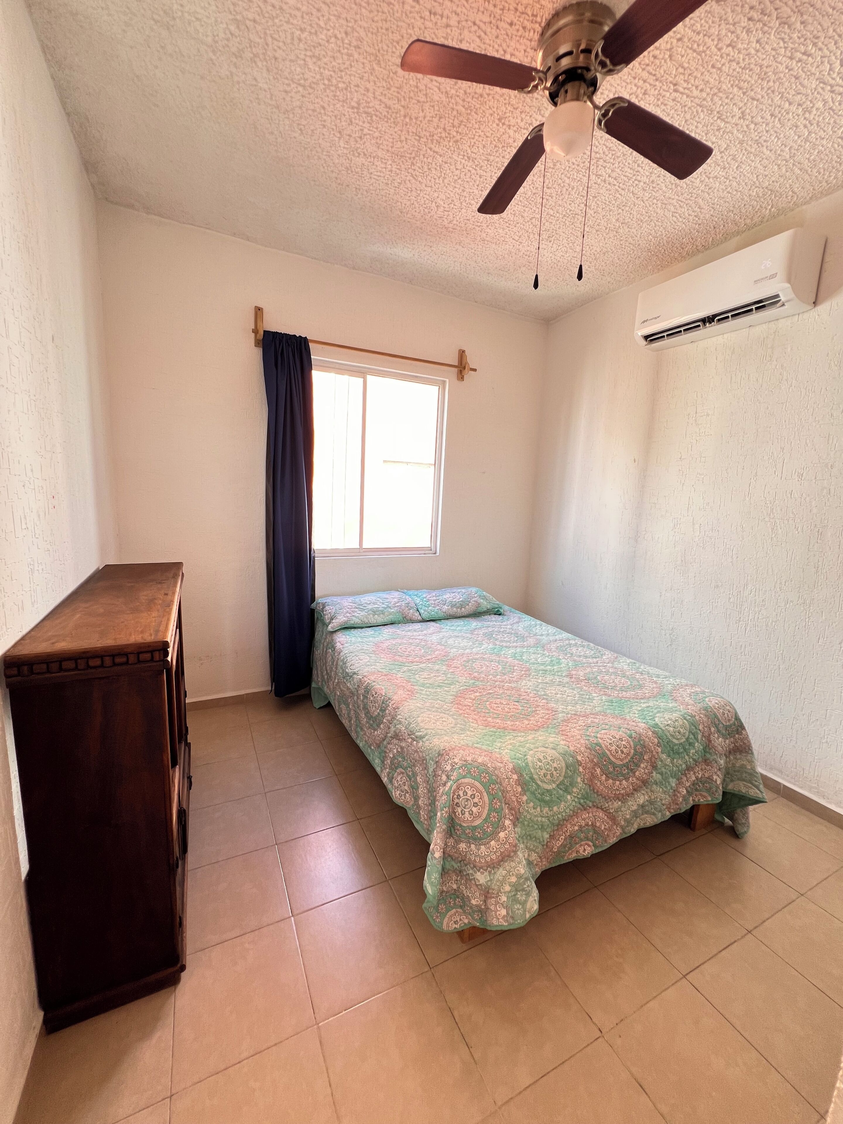 Une chambre à coucher comprenant un lit, un ventilateur de plafond, une fenêtre avec des rideaux, une commode en bois et un appareil de climatisation.
