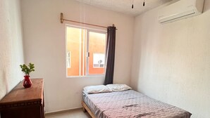 A bedroom with a bed, a ceiling fan, a window with curtains, a small table with a plant, and an air conditioning unit. - Relax in paradise (Playa del Carmen)