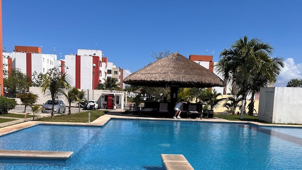 A swimming pool with a thatched-roof structure, surrounded by buildings and palm trees. - Relax in paradise (Playa del Carmen)