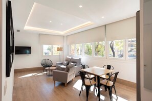 Un salon moderne comprenant un canapé, une table à manger et des chaises, un téléviseur à écran plat fixé au mur et de grandes fenêtres offrant une vue sur l’extérieur.