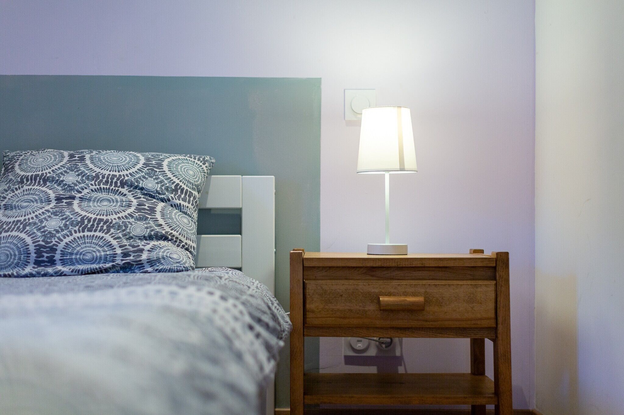 A bed with patterned bedding, a bedside table with a lamp, and a wall-mounted light switch.