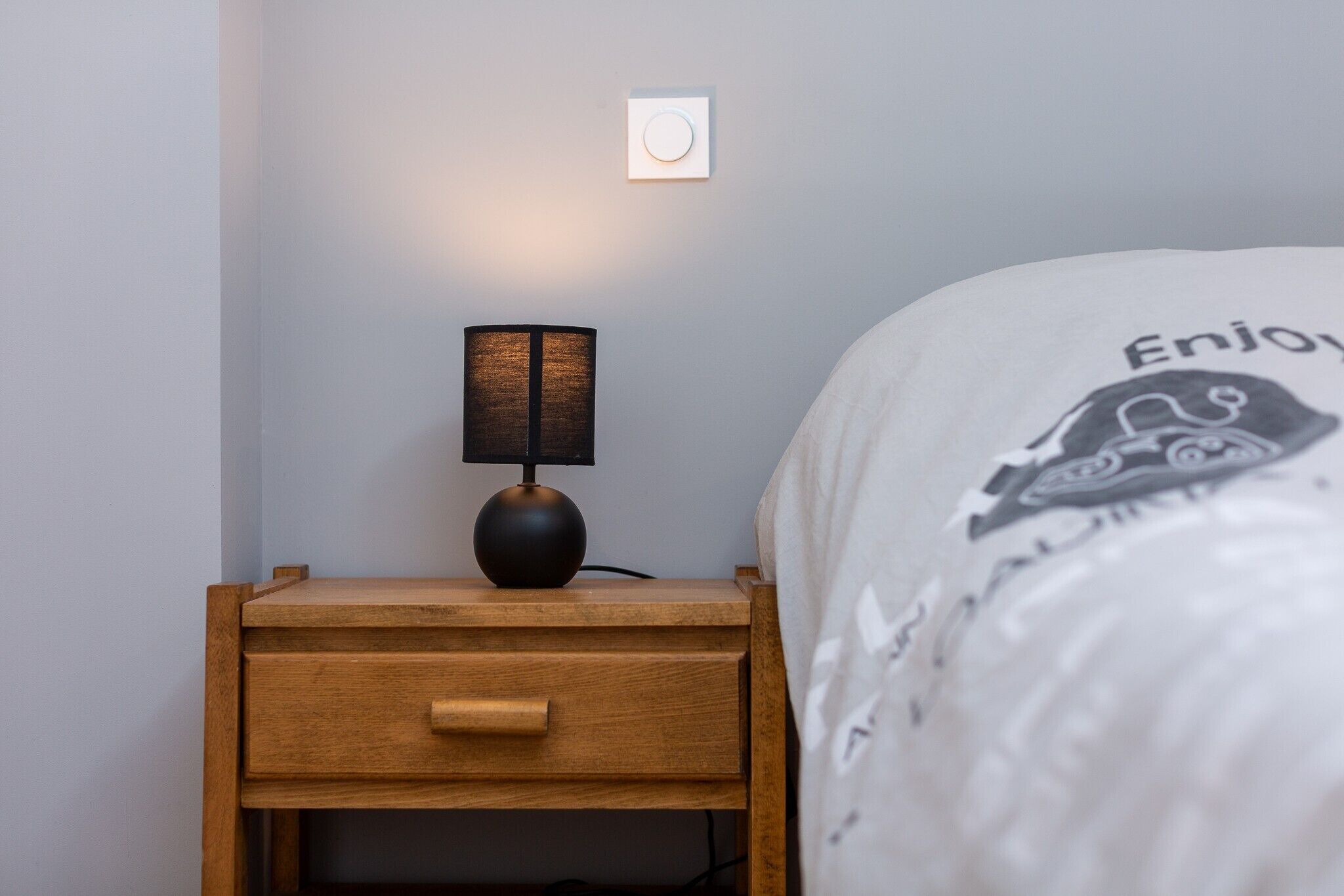 A wooden bedside table with a lamp, a drawer, and a bed with a white comforter.
