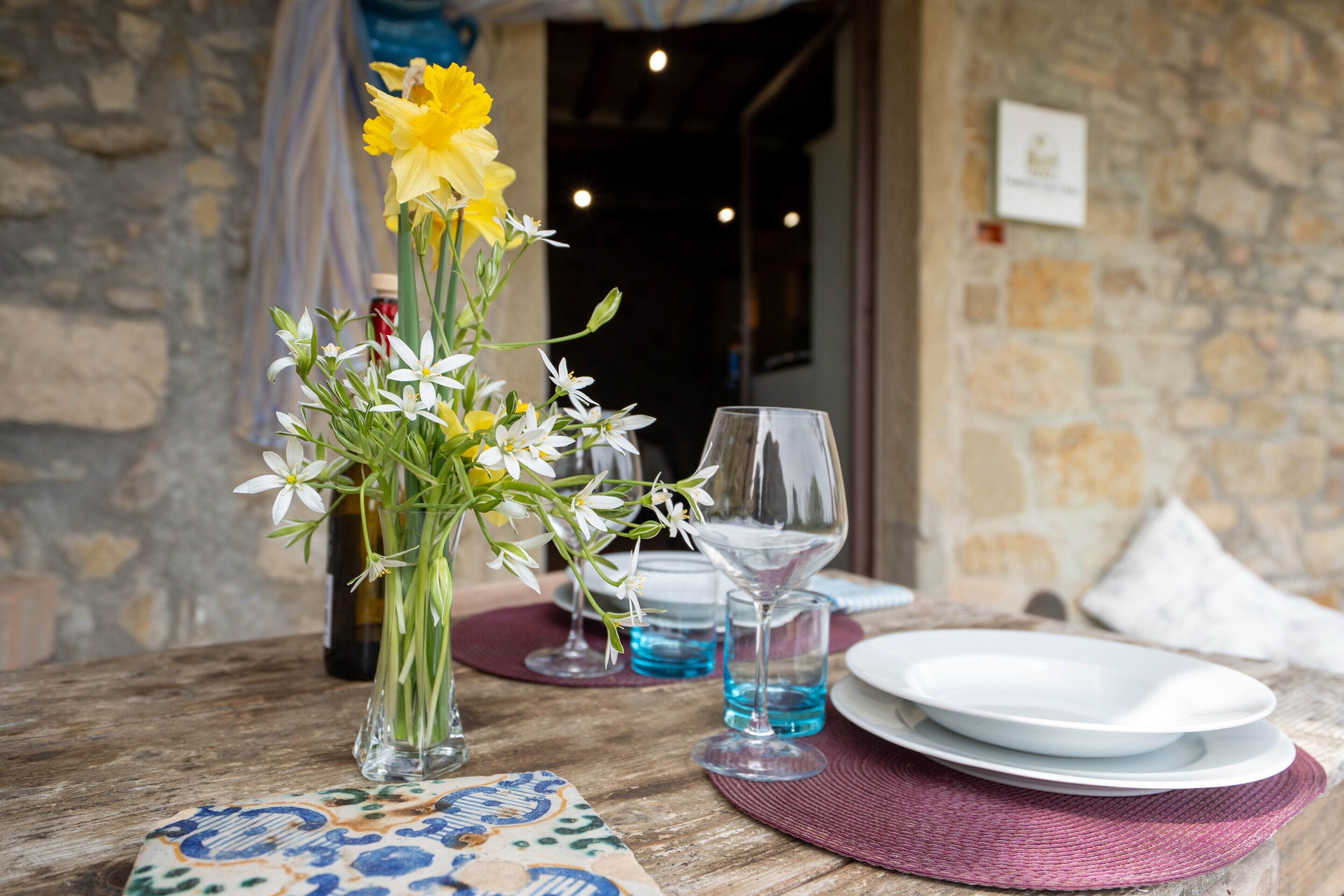 A table with a vase of flowers, wine glasses, and plates, set against a stone wall.