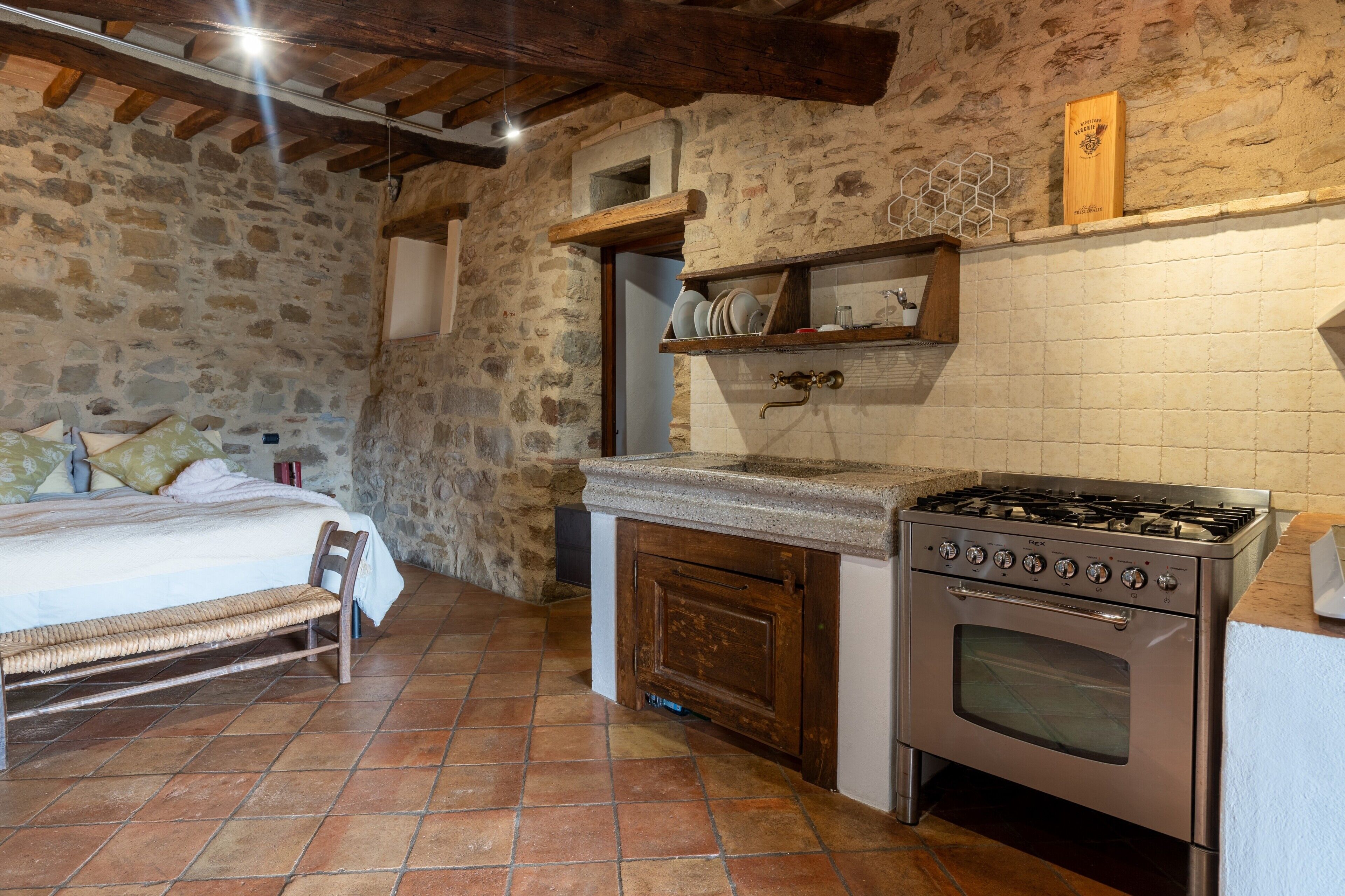 A rustic kitchen with a stone wall, wooden beams, a bed with a bench, and a stove.