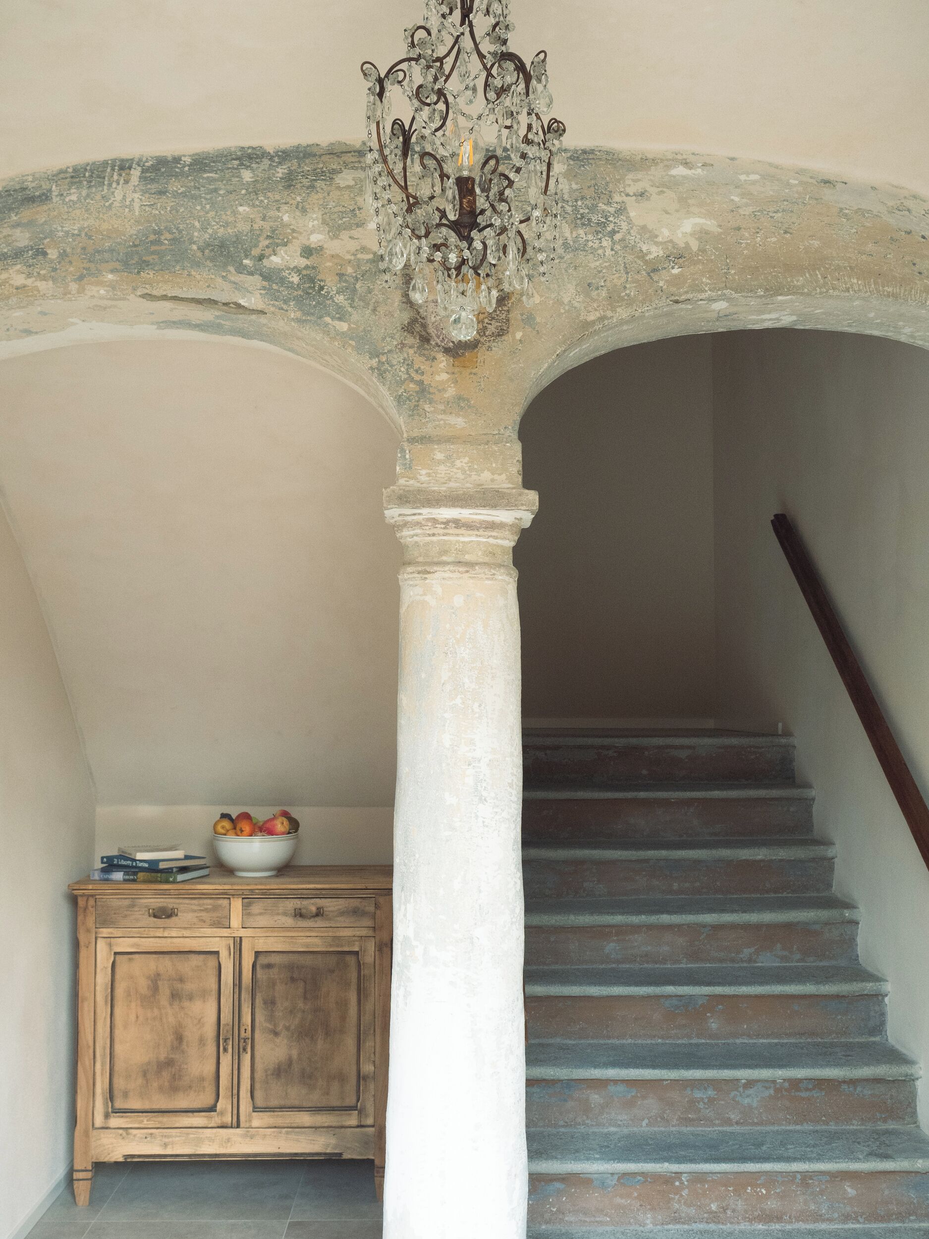Un escalier avec une rampe en bois, une colonne blanche et un saladier de fruits posé sur un meuble en bois.