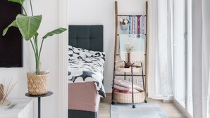 Ein Schlafzimmer mit Bett, Schreibtisch, einer großen Pflanze, einem Kleiderschrank und einem Fenster mit Vorhängen.