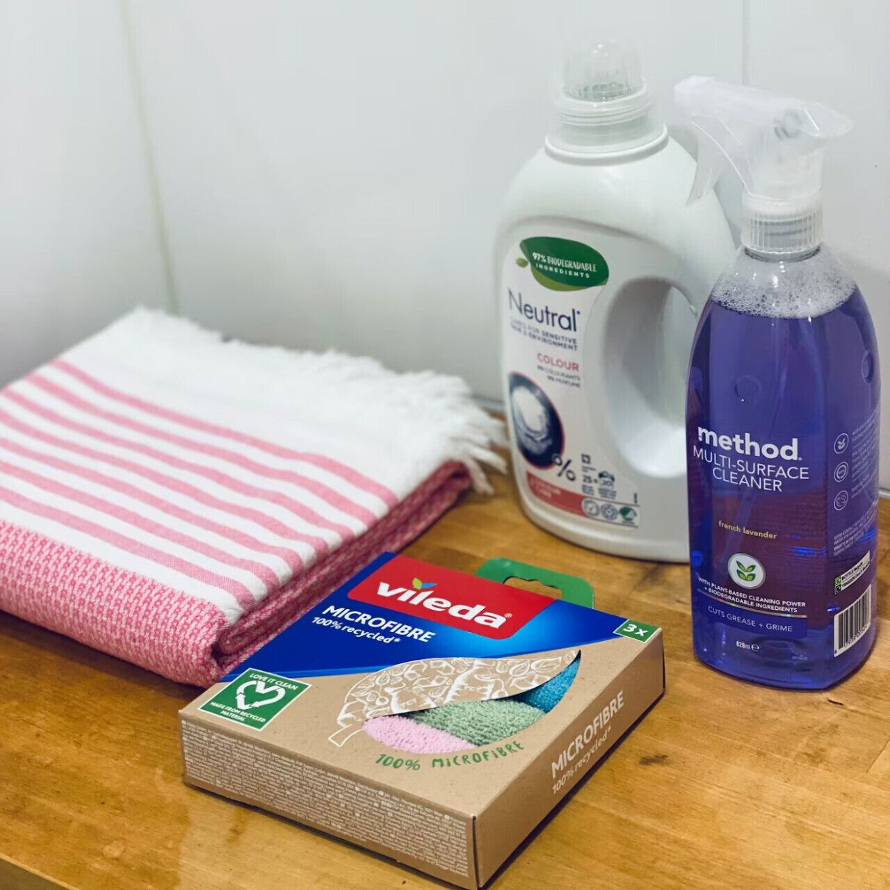 Una superficie de madera con una toalla blanca, una botella de detergente Neutral, una botella de limpiador multiuso Method y una caja de paños de microfibra Vileda.