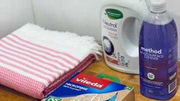 Una superficie de madera con una toalla blanca, una botella de detergente Neutral, una botella de limpiador multiuso Method y una caja de paños de microfibra Vileda.