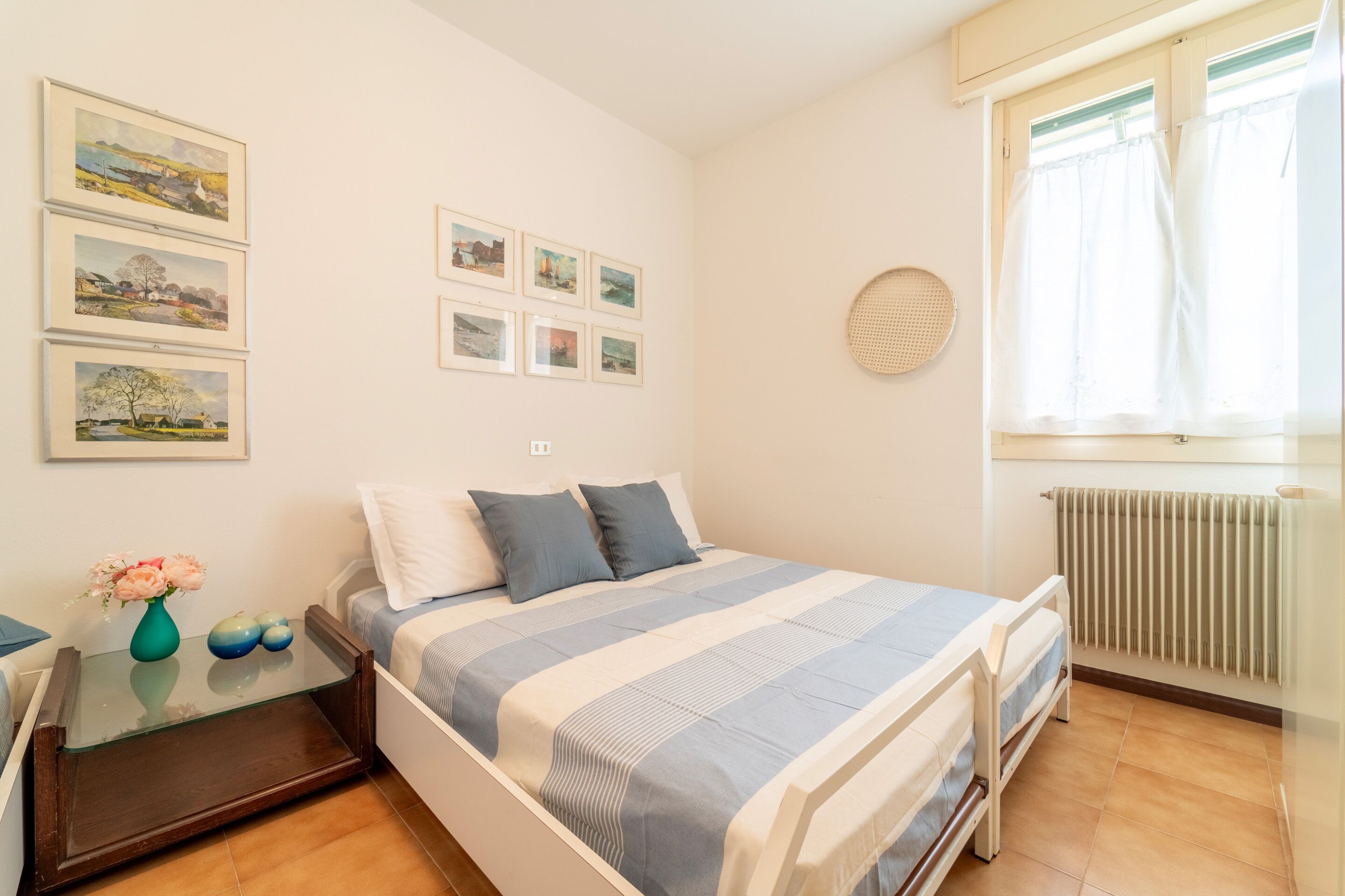 Un dormitorio con cama, mesitas de noche, cómoda, ventana con cortinas y cuadros en la pared.