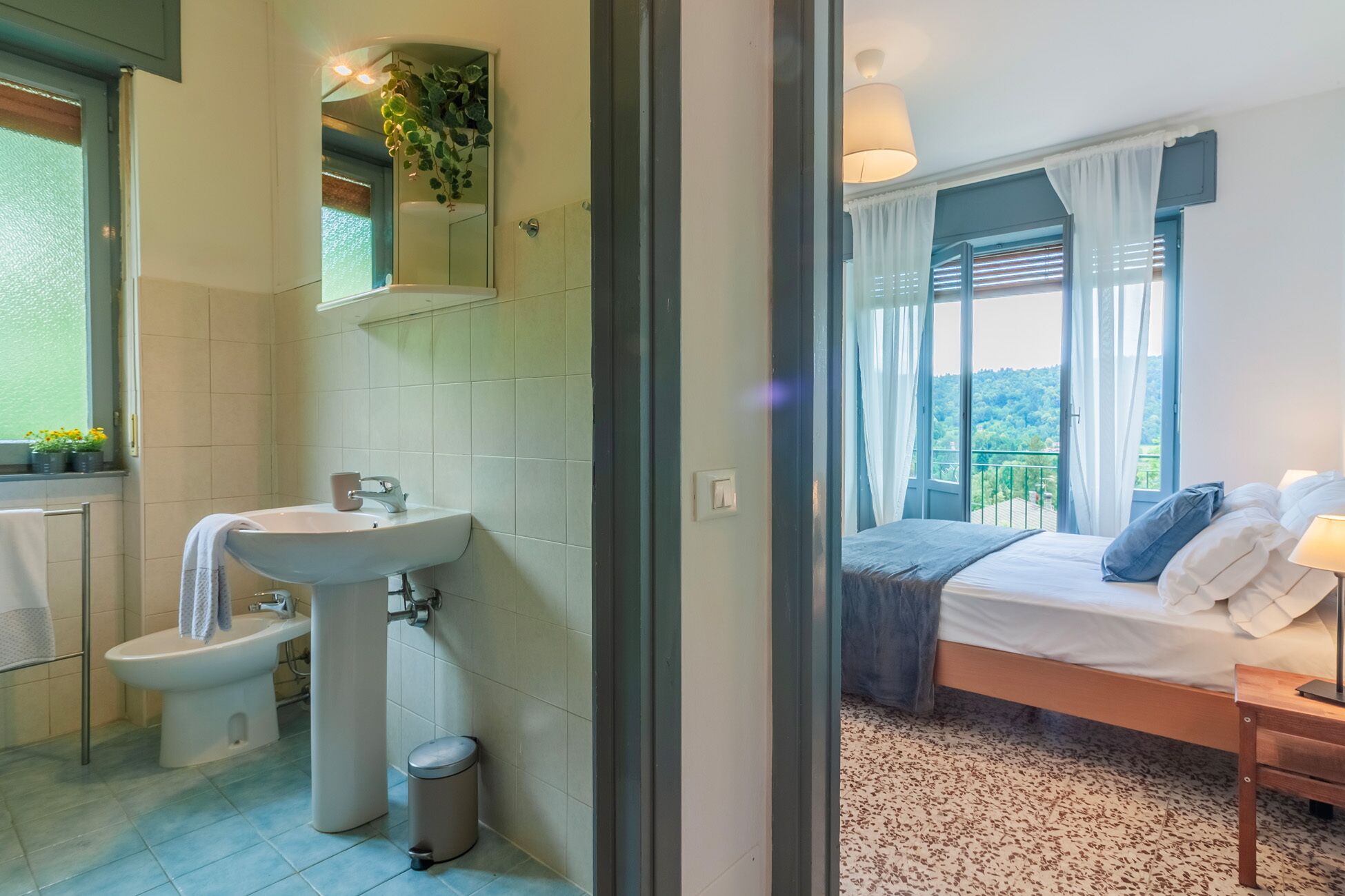 A bathroom with a sink, toilet, and a small trash bin. The room has a window with curtains and a view of greenery outside.