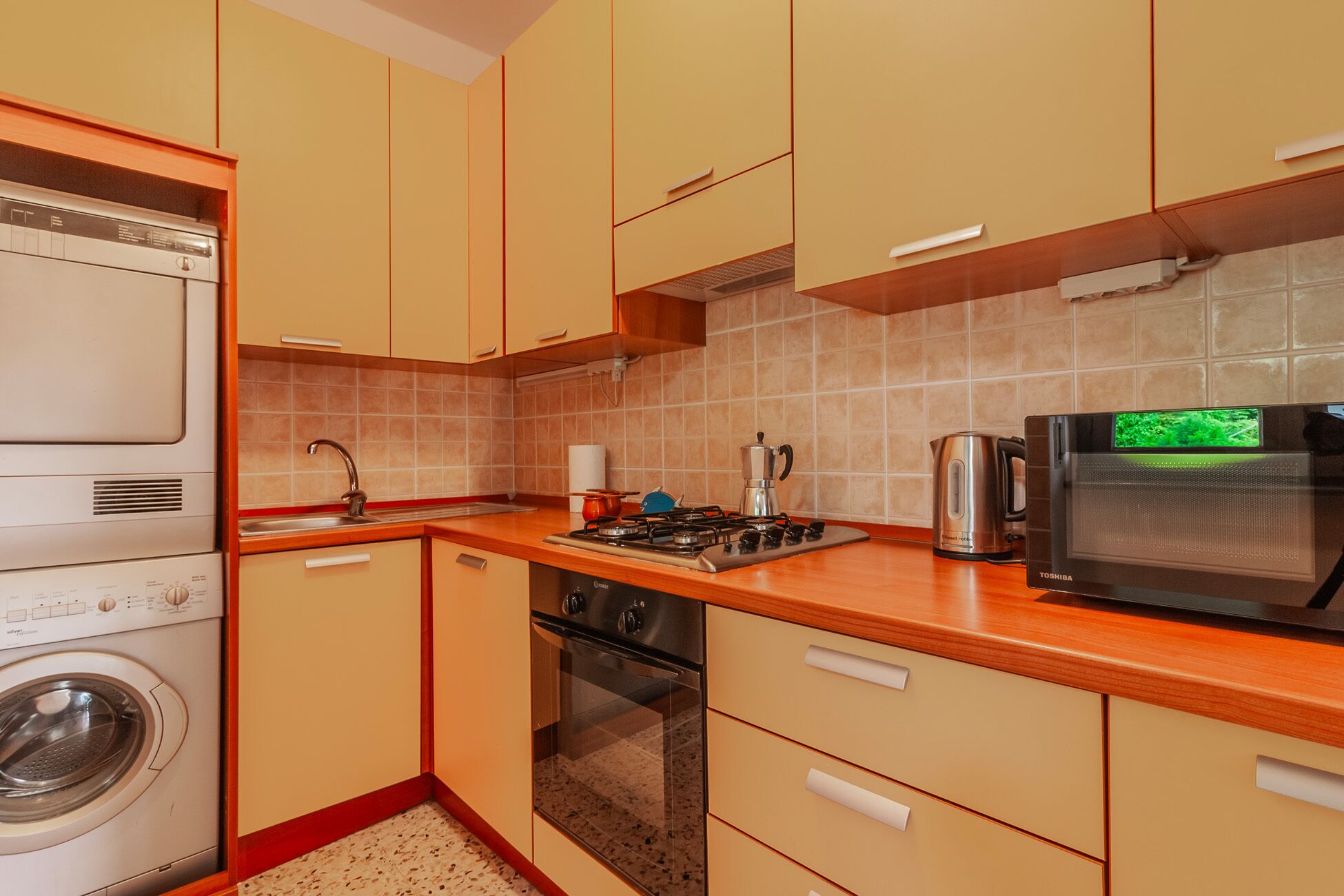 A compact kitchen with a washing machine, oven, microwave, and a kettle.