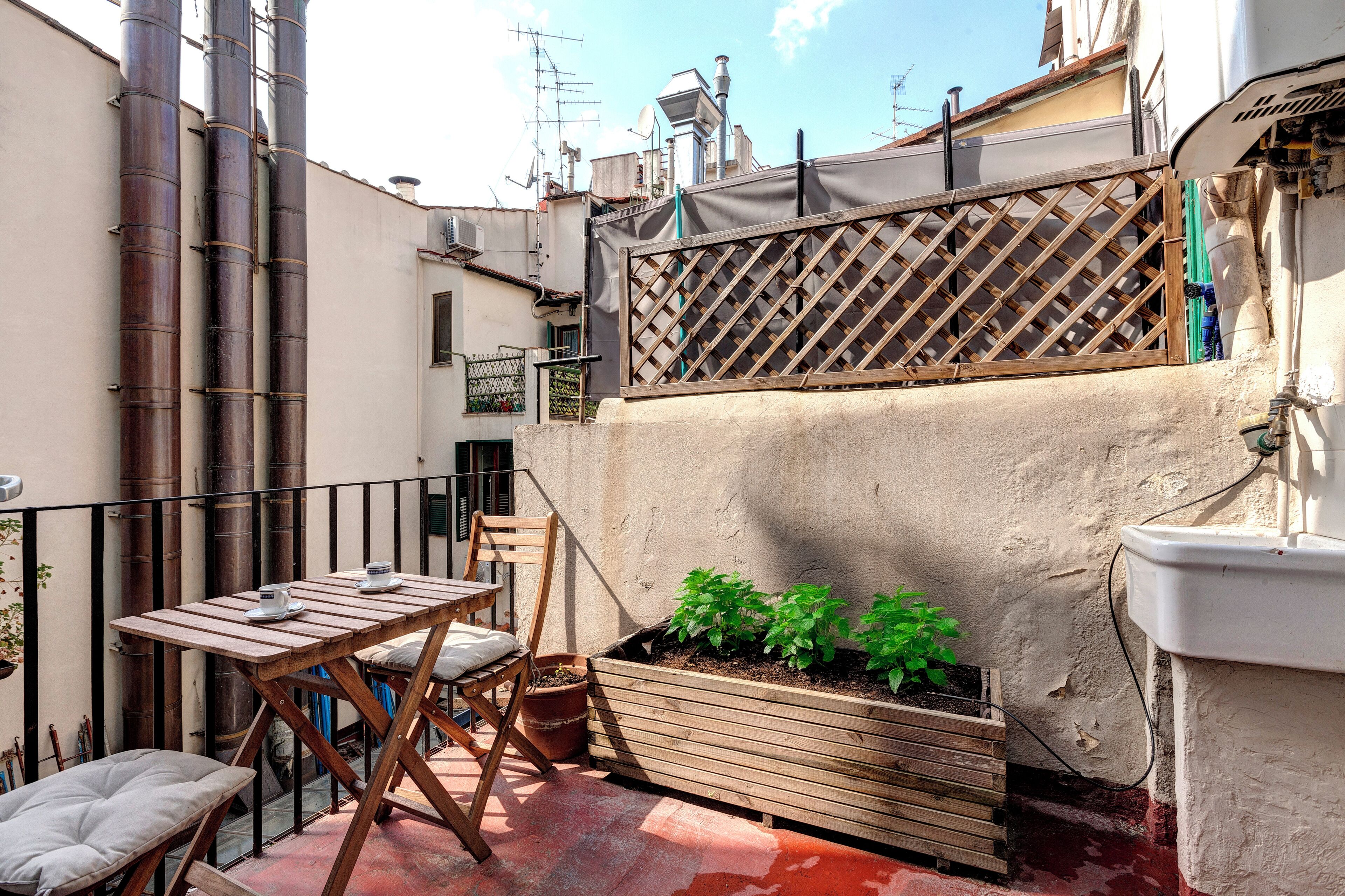 A small outdoor patio with a wooden table and chairs, a planter box with plants, and a metal fence.