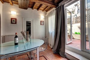 Een glazen tafel met een fles wijn, transparante stoelen, een witte deur en uitzicht op een binnenplaats door een raam.