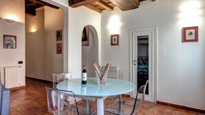Uma área de refeições com mesa de vidro, cadeiras, uma garrafa de vinho e um vaso com galhos.