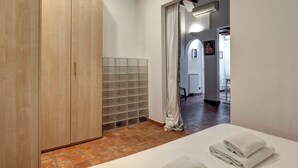 Quarto de hotel com guarda-roupa de madeira, chão em mosaico, cama com toalhas dobradas e vista para uma sala de jantar através de uma porta aberta.
