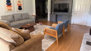A living room with a grey sofa, brown couch, wooden chairs, a coffee table, and a TV on a grey cabinet. - Fully Equipped Brandon Home — Comfortable & Convenient off Hwy 471 (Brandon)