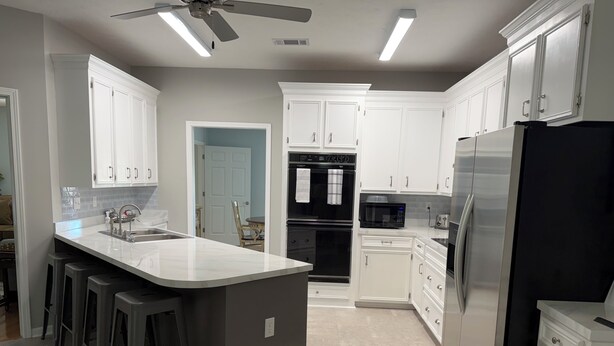A modern kitchen with white cabinets, a large island with bar stools, a black refrigerator, and a ceiling fan. - Fully Equipped Brandon Home — Comfortable & Convenient off Hwy 471 (Brandon)