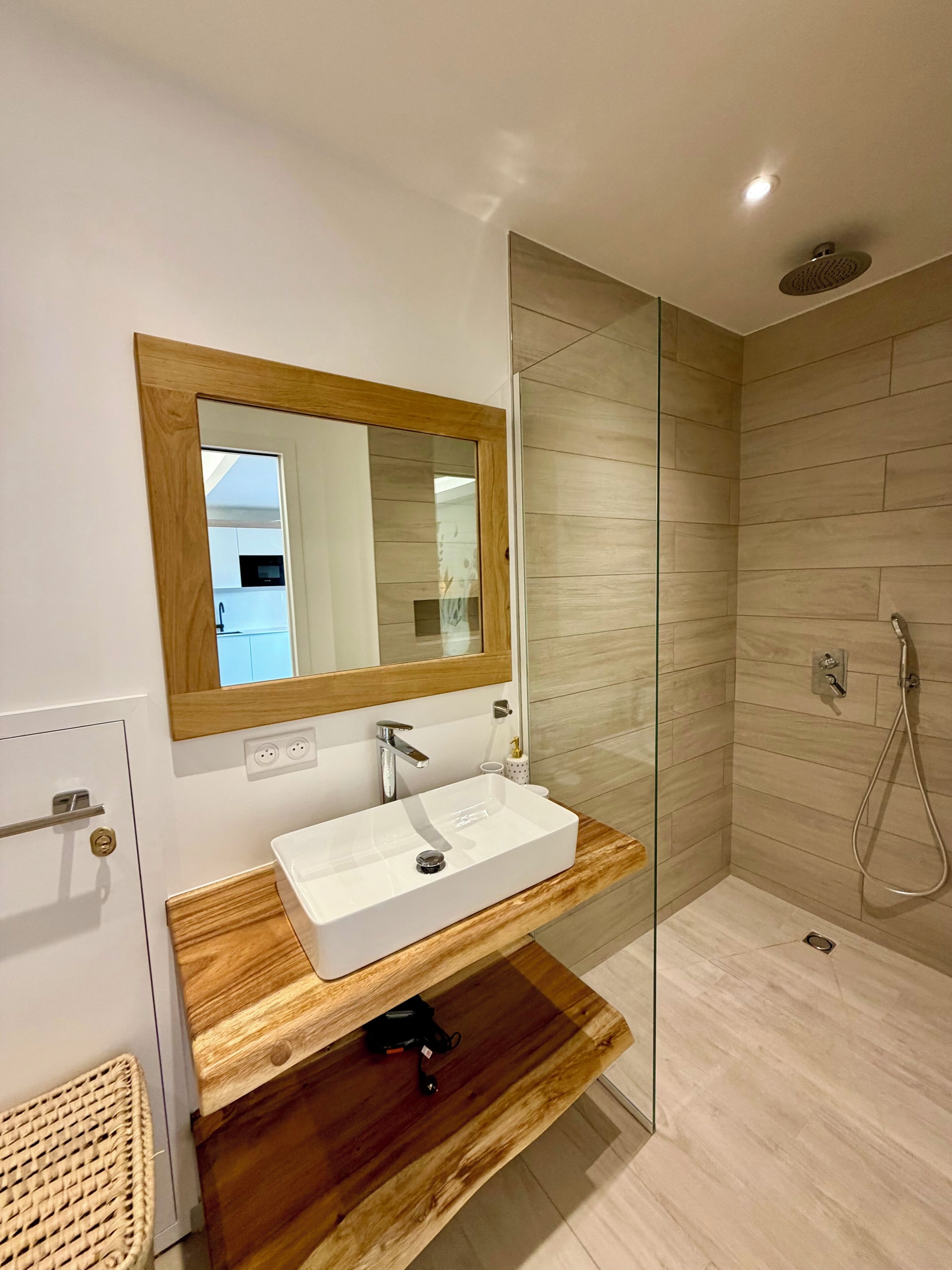 Un baño moderno con tocador de madera, un lavabo blanco, un espejo con marco de madera y una zona de ducha.