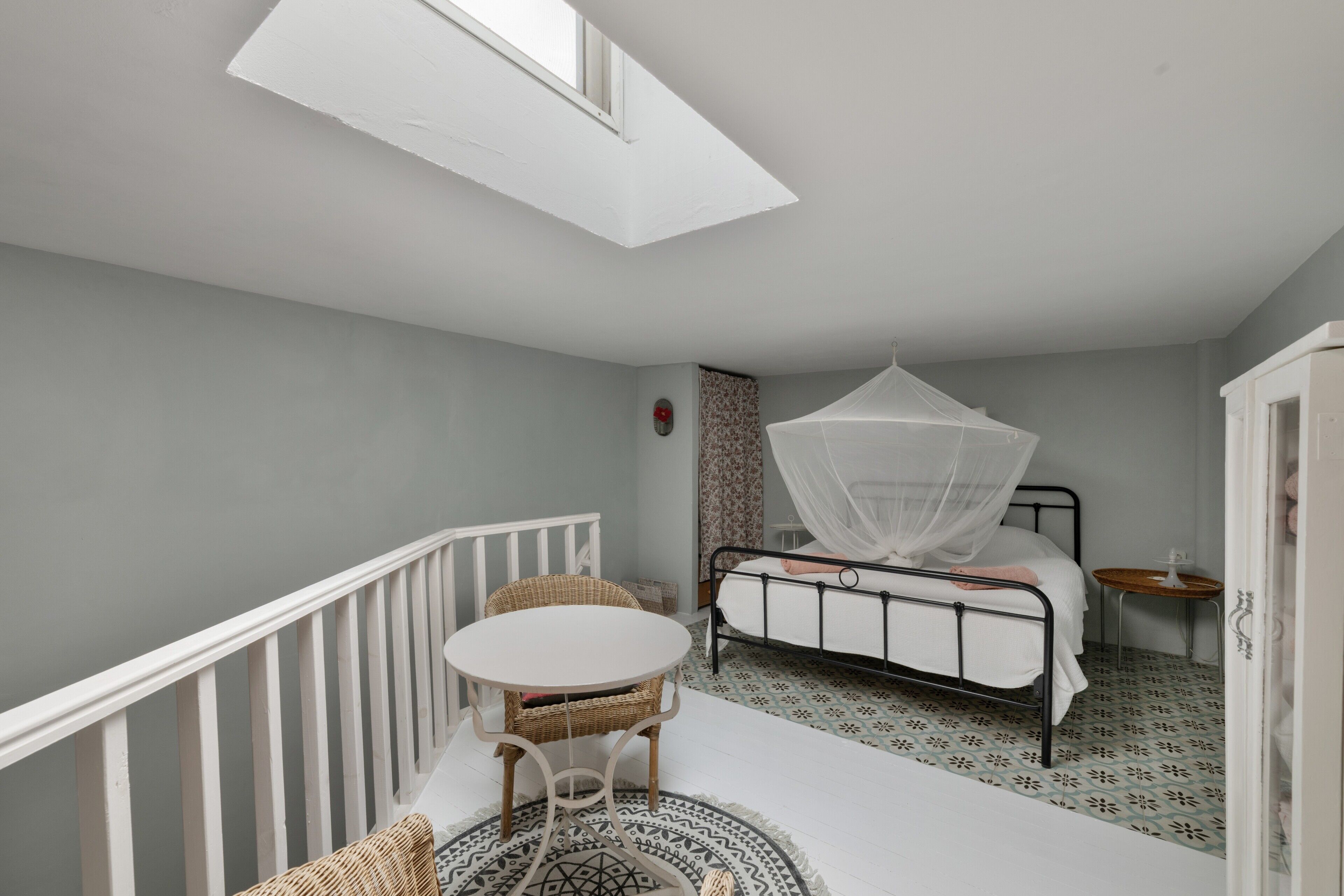 Een slaapkamer met een bed, nachtkastje, stoel, klein tafeltje, raam en een balkonhek.