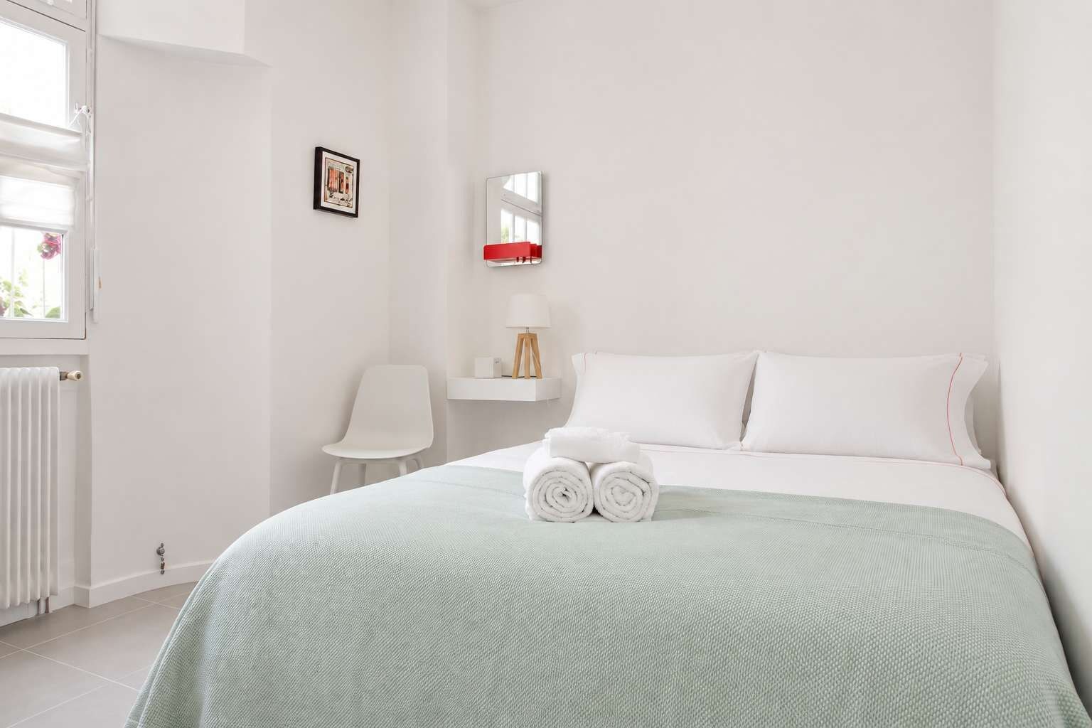 A hotel room with a bed, a chair, a bedside table, a radiator, and a window.