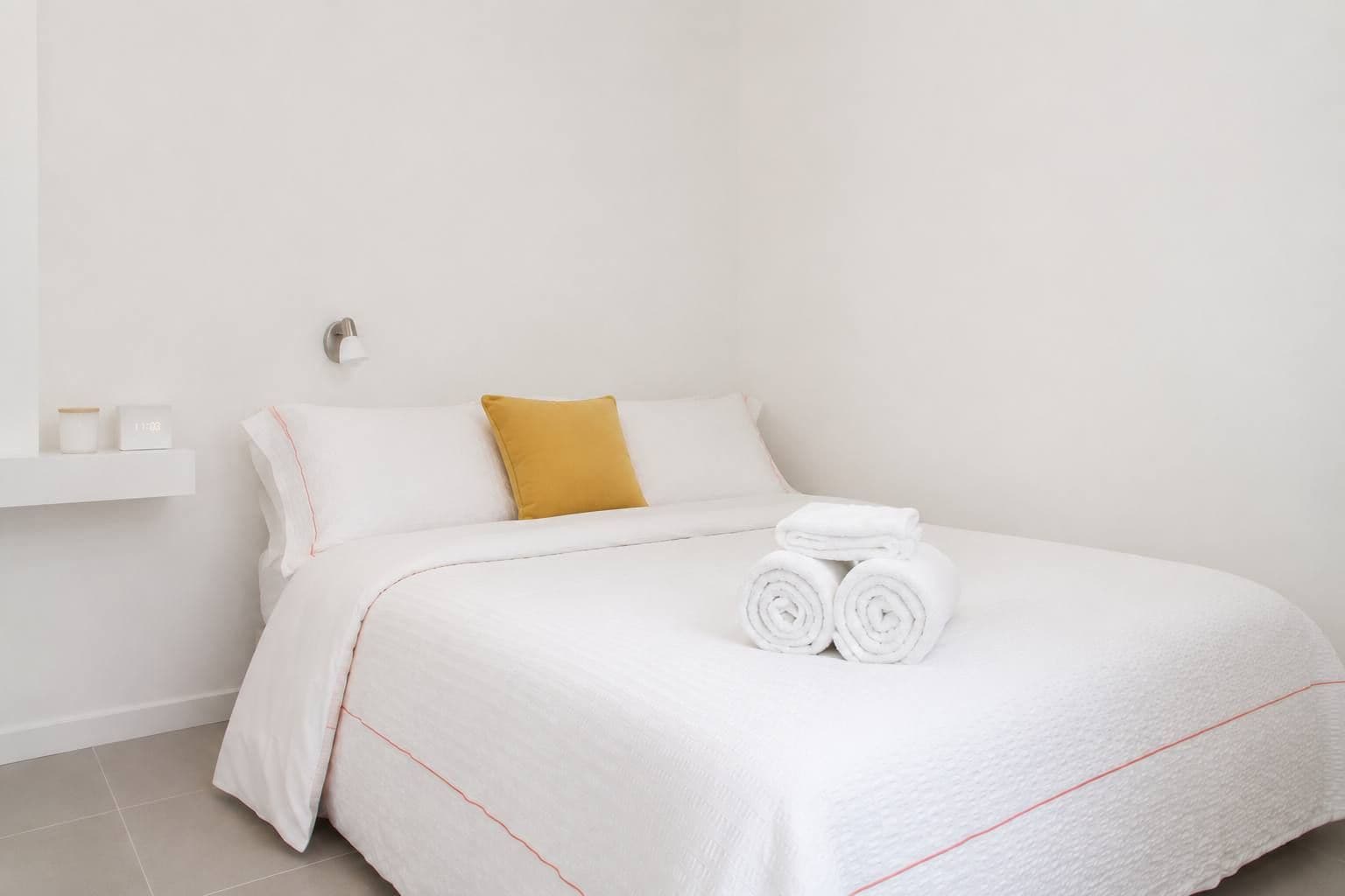 A neatly made bed with white linens and a single rolled towel, a mustard-colored pillow, and a small white shelf with two items.