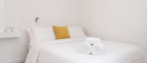 A neatly made bed with white linens and a single rolled towel, a mustard-colored pillow, and a small white shelf with two items.