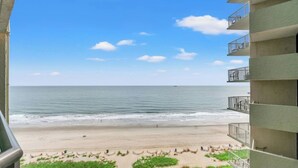 Une vue sur la plage depuis un balcon donnant sur une étendue de sable, avec une mer calme, un ciel dégagé et un immeuble doté de nombreux balcons.