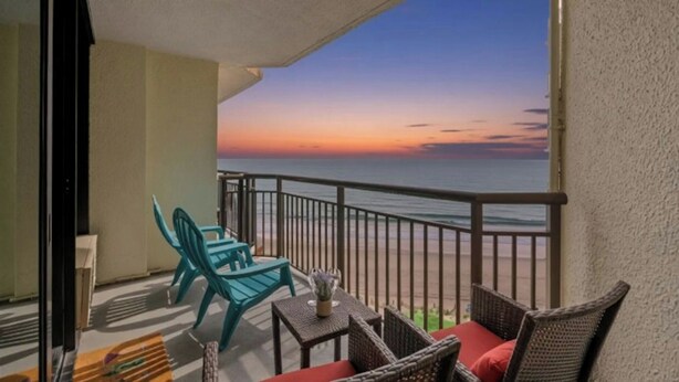 Un balcon avec vue sur l’océan au coucher du soleil, des meubles en osier et une petite table sur laquelle est posé un vase.