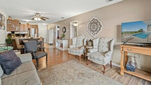 A living room with a sofa, armchairs, a TV, and a dining area with a kitchen in the background. - Crescent Villas 302- 2 Bedroom Condo- Pool (North Myrtle Beach)