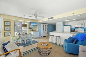 A spacious living area with a dining set, a blue sofa, a kitchen with white cabinets, and a ceiling fan. - Buena Vista 401-Oceanfront 3BR Condo- Cherry Grove (North Myrtle Beach)