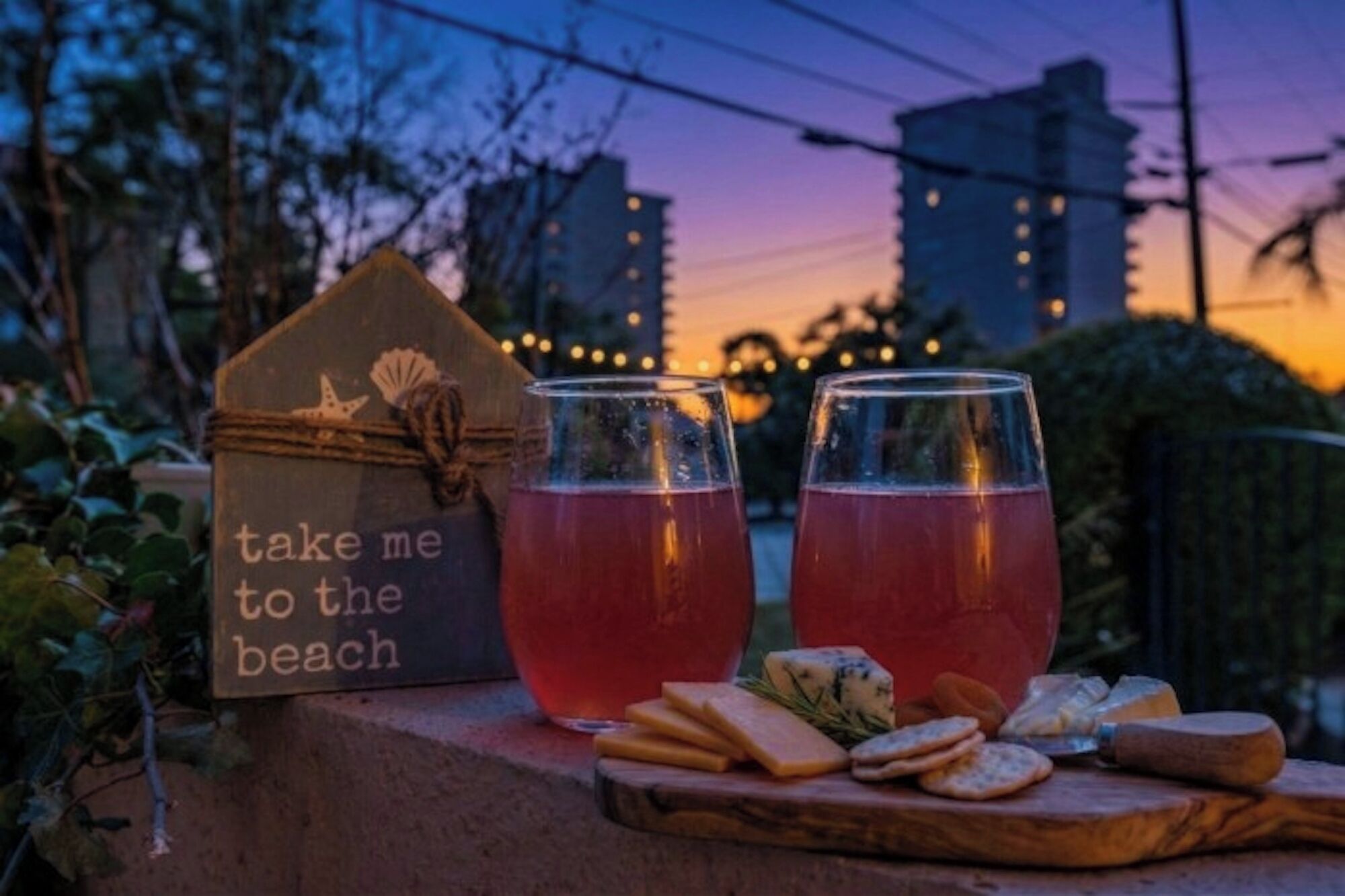 A wooden sign with a message, two glasses of red beverage, and a selection of cheese and crackers on a wooden board.