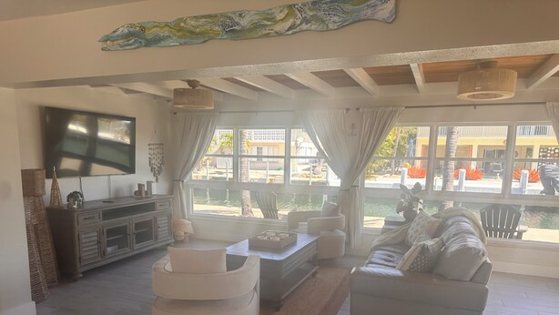 A spacious living room with a large TV, comfortable seating, a dining area, and a view of a pool. - Ocean Breeze Retreat/ Elegant 3/3 in Marathon/ Coastal Luxury Living (Marathon)