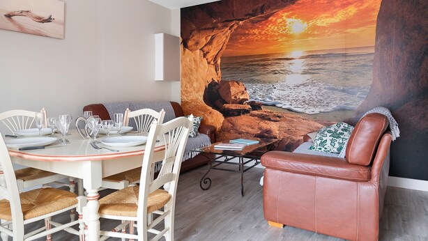 Un'area pranzo con tavolo apparecchiato per quattro, una poltrona e un grande quadro raffigurante un tramonto sul mare.