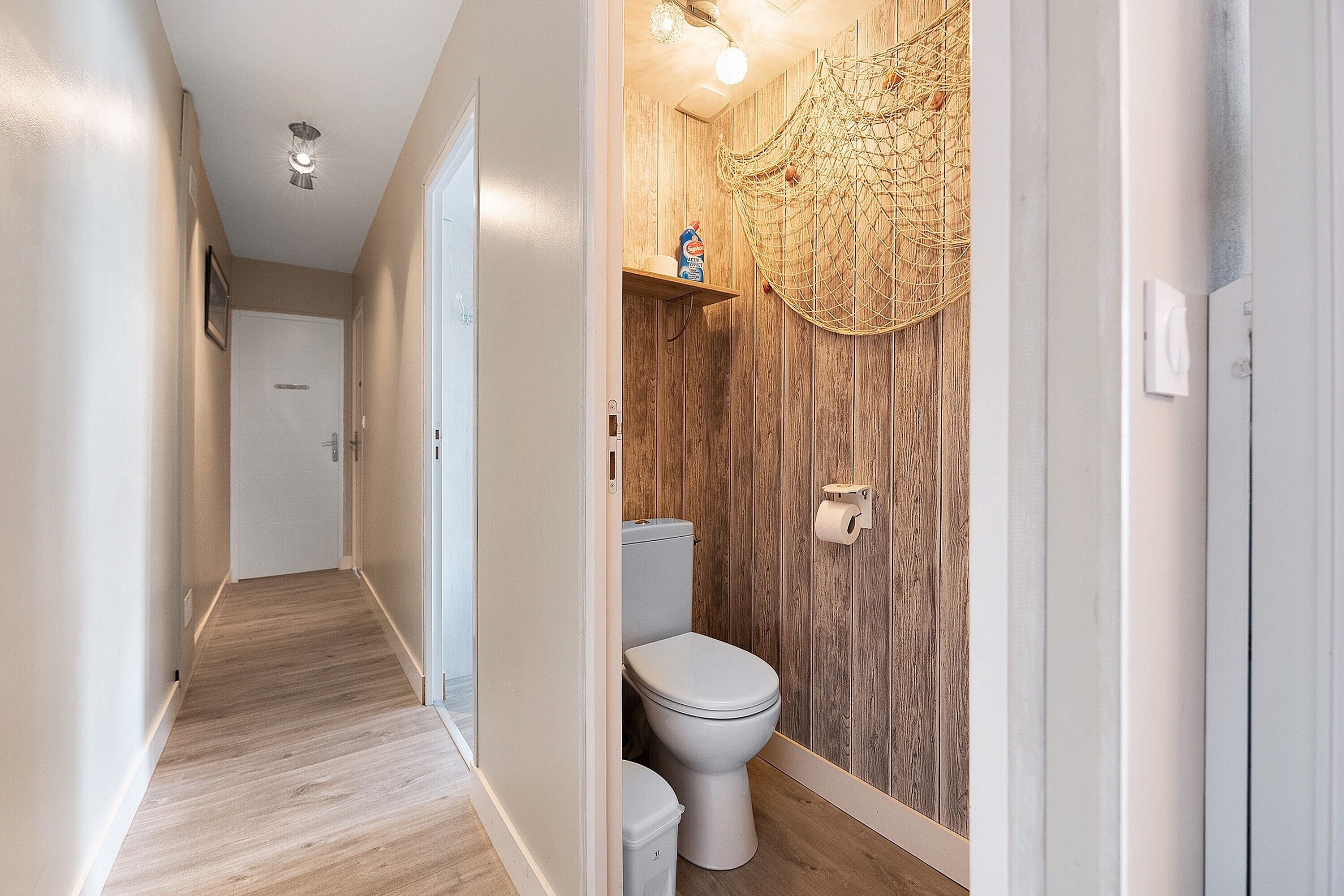 A hallway leading to a bathroom with a wooden wall, a toilet, and a shelf with a bottle.