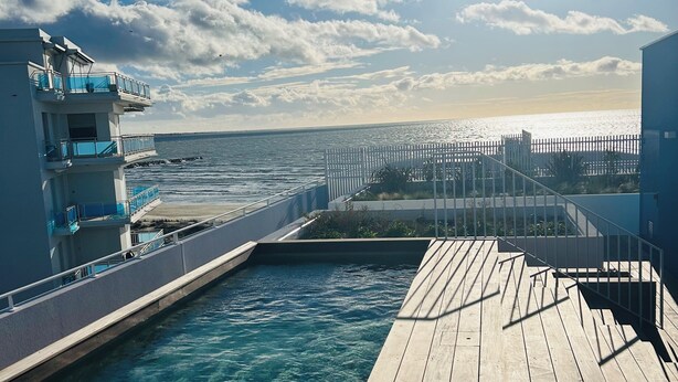 Una terrassa de fusta amb aigua cristal·lina que condueix a una piscina, un edifici blanc amb balcons, i vista al mar.