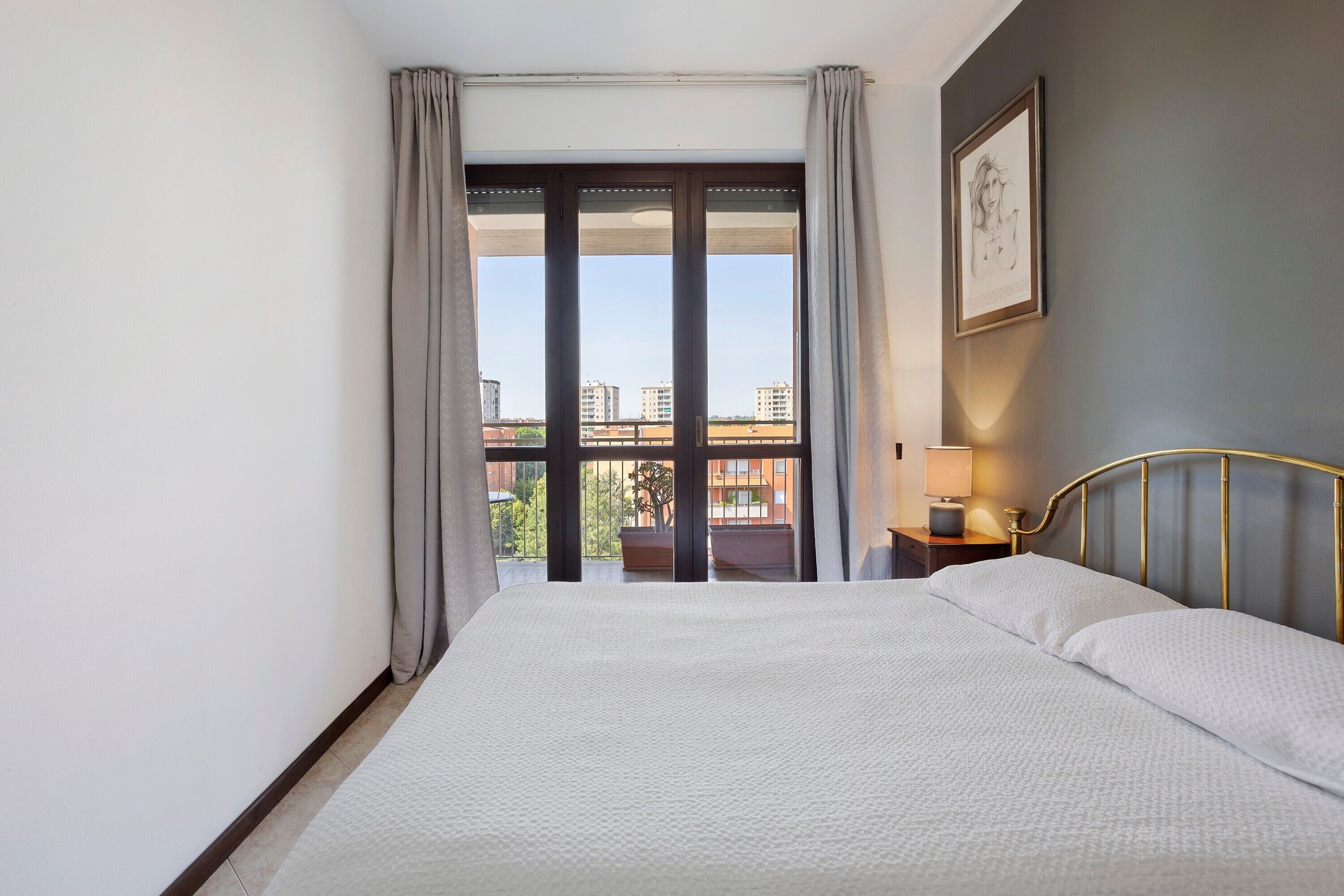 A hotel room with a large bed, a nightstand with a lamp, a framed picture on the wall, and a view of buildings through the window.