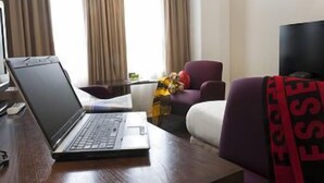 Une chambre d’hôtel avec un ordinateur portable sur un bureau, un fauteuil violet, un lit recouvert d’une couette blanche et un téléviseur placé à droite.