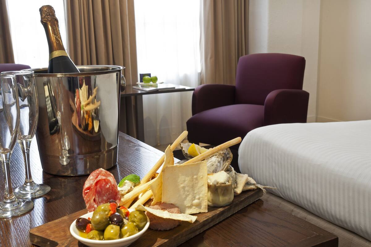 A hotel room with a bed, a wooden table with a bottle of champagne, glasses, and a board with olives, cheese, and meat.