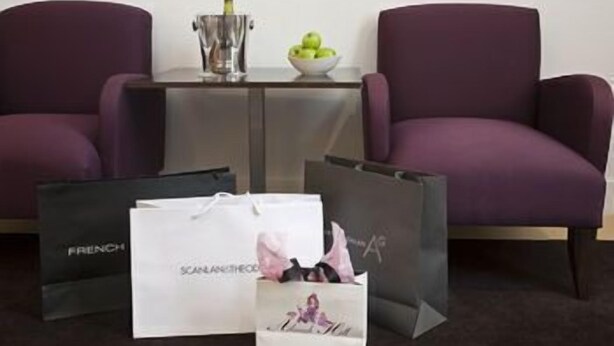 Une pièce avec deux fauteuils violets, une petite table sur laquelle se trouvent une bouteille et un saladier rempli de fruits, et trois sacs de courses.