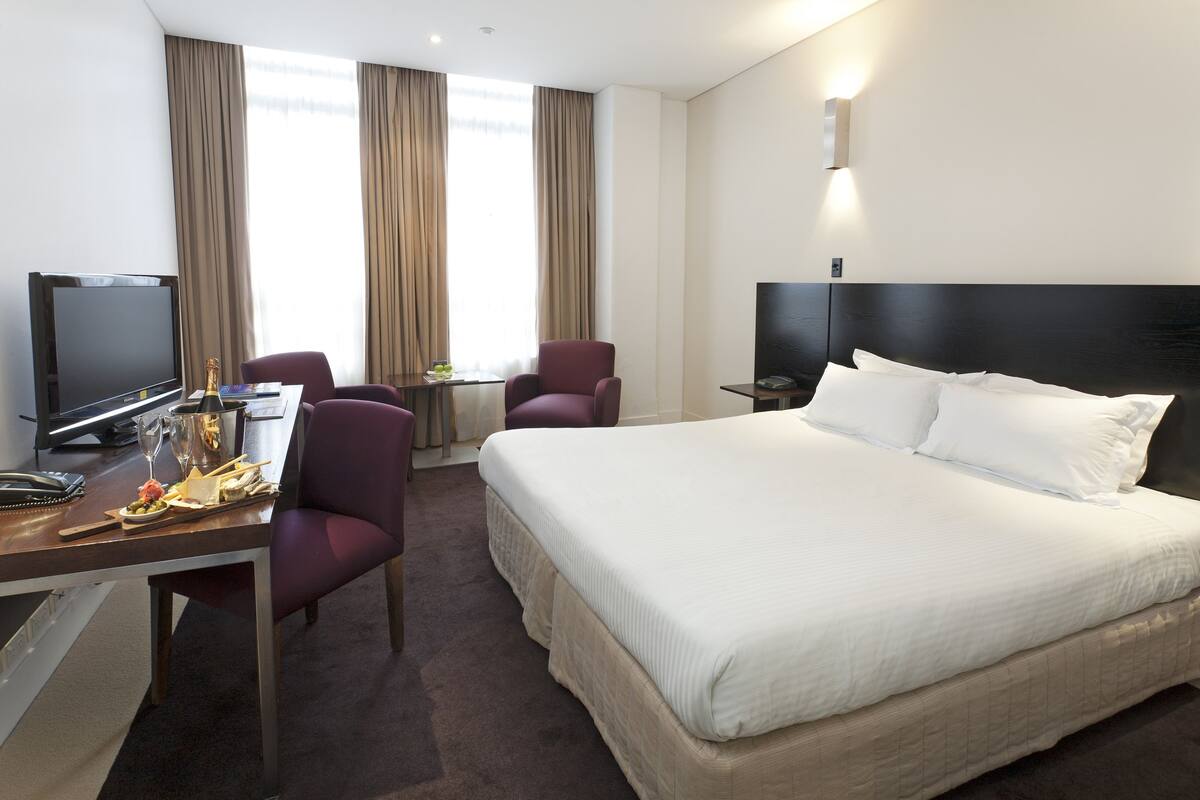 A hotel room with a large bed, a desk with a computer and a chair, a television, and a window with curtains.