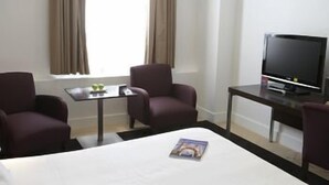 Une chambre d’hôtel comprenant un lit, deux fauteuils, une petite table avec une télécommande, un téléviseur et une fenêtre avec des rideaux.
