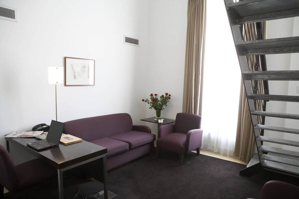 A room with a desk, purple sofa, chair, and a small table with a vase of flowers.