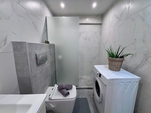 A compact bathroom with a toilet, a washing machine, and a shower stall. - Bright 1BR Apartment 5 min to Ladadika (Thessaloniki)
