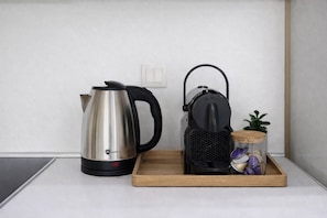 Una encimera de cocina con un hervidor de acero inoxidable, un hervidor eléctrico negro y una bandeja de madera que contiene un tarro de vidrio con una planta y algunos objetos morados.