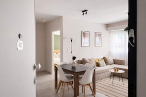 A modern living room with a sofa, a dining table, and chairs. There is a door with a number plate, a window with curtains, and a wall-mounted light fixture. - Bright 1BR Apartment 5 min to Ladadika (Thessaloniki)