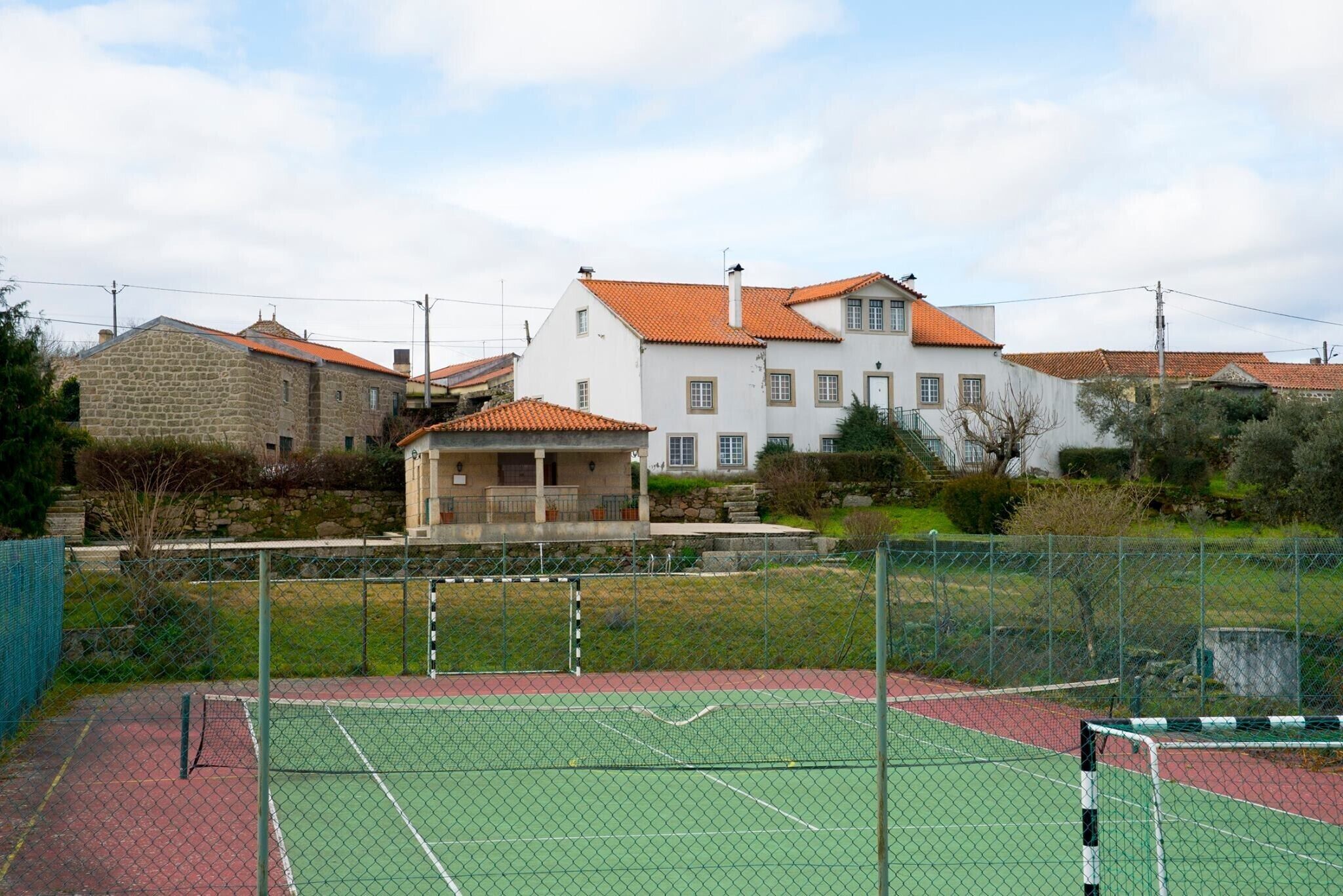 Una pista de tennis amb una xarxa, envoltada per una tanca, i un edifici amb teulada vermella al fons.