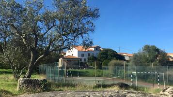 Un court de tennis entouré d’une clôture, avec un poteau de but, un grand arbre et un bâtiment avec un toit rouge en arrière-plan.