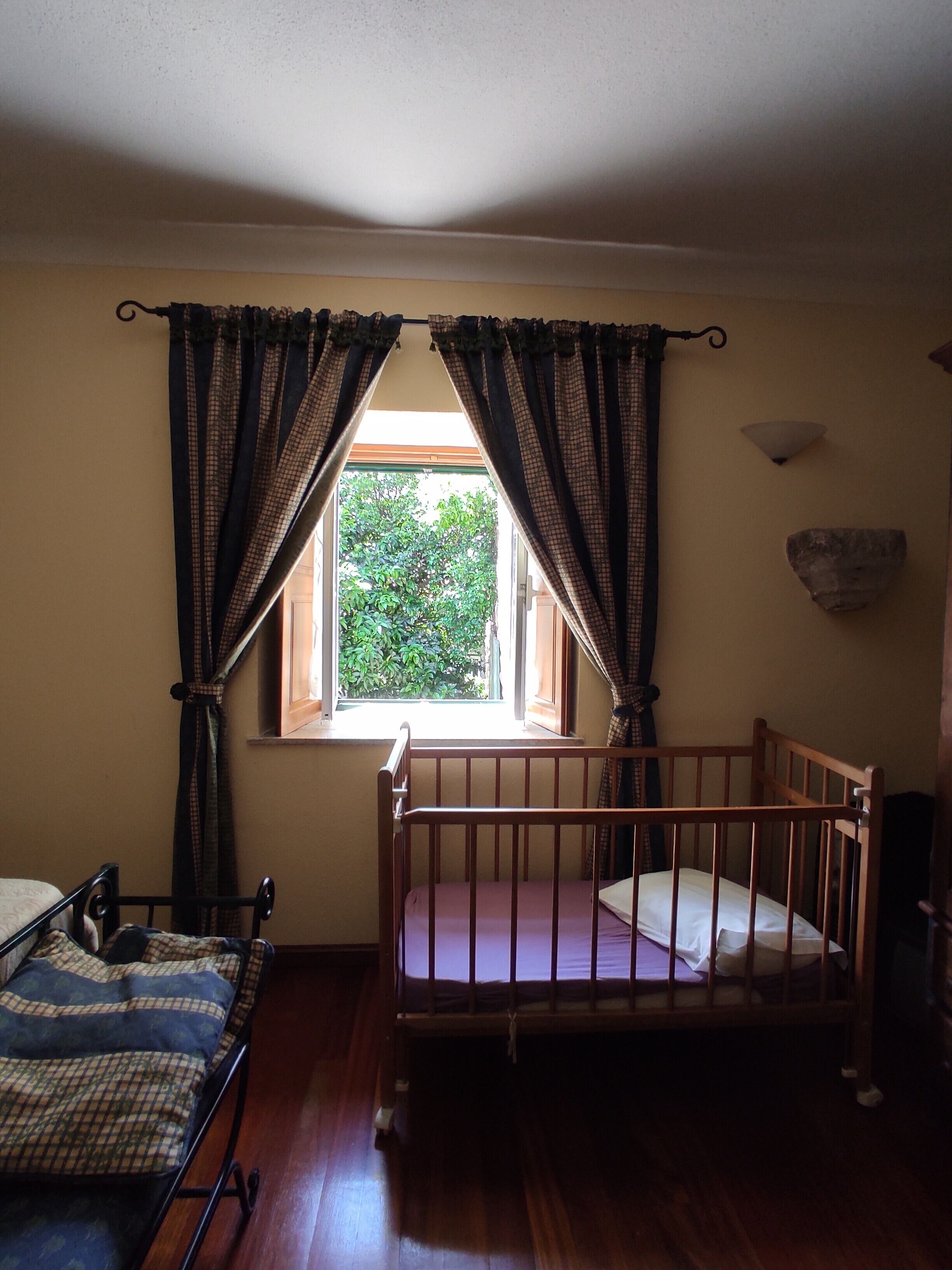 A room with a wooden crib, a window with curtains, a wall-mounted light fixture, and a radiator.