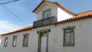 A two-story building with a white facade, a red-tiled roof, and a black wrought-iron balcony.