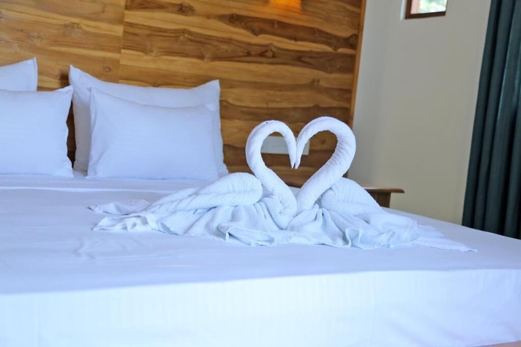 Cama com roupa de cama e almofadas brancas, cabeceira de madeira e arranjo de toalhas em formato de coração.